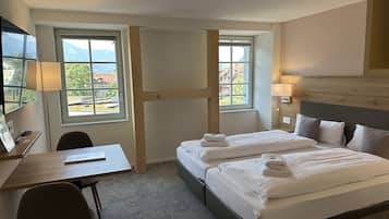 Et hotelværelse med to senge, et skrivebord, en stol og udsigt til det fri gennem store vinduer.