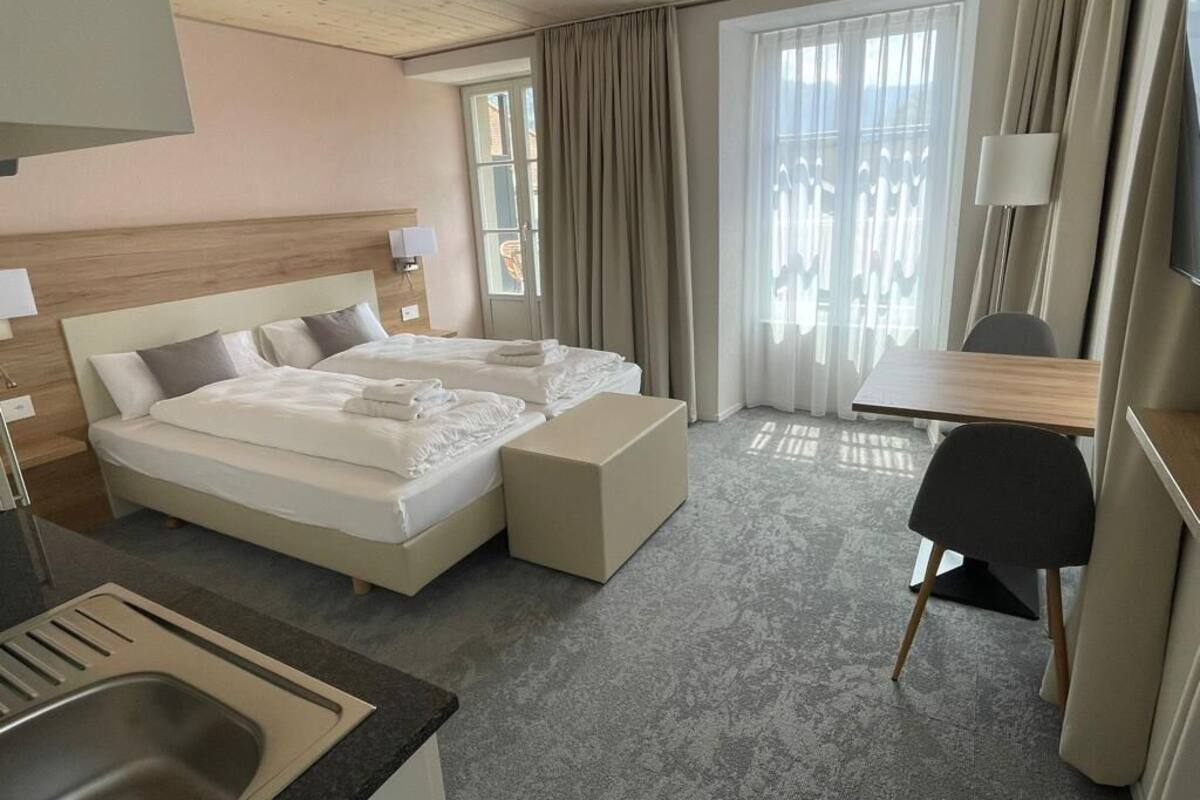 A hotel room with a double bed, a desk, a chair, a small kitchenette, and a large window with curtains.