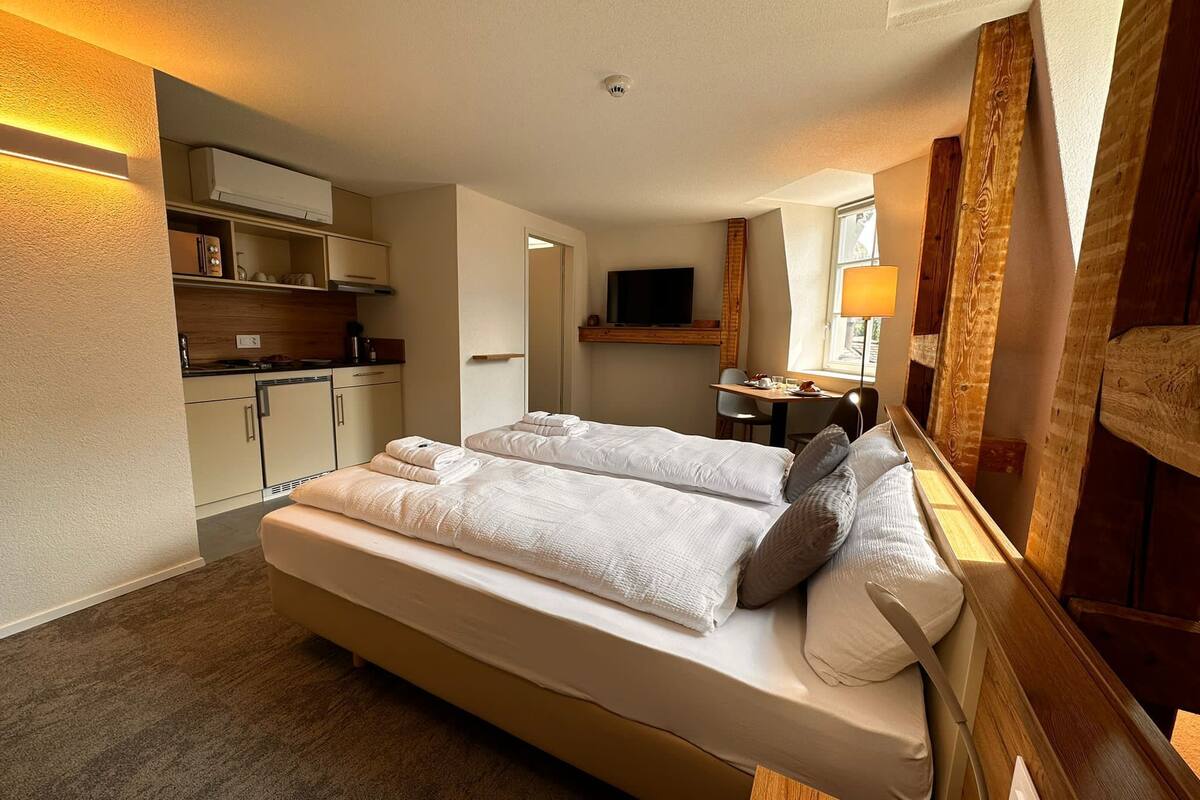 A hotel room with a large bed, a desk, a chair, a TV, and a kitchenette.