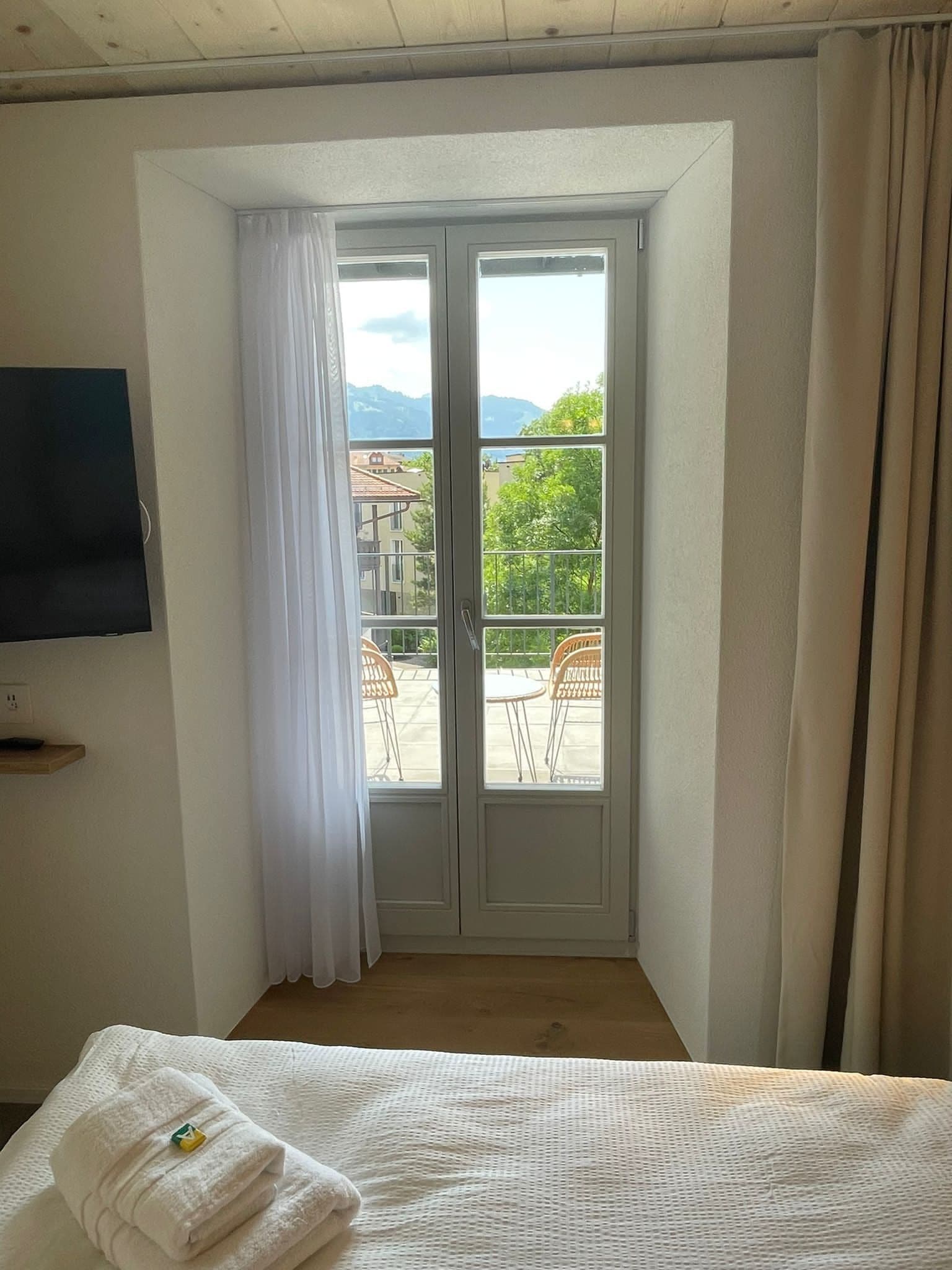 A hotel room with a bed, a TV mounted on the wall, a balcony with chairs, and a view of buildings and mountains.