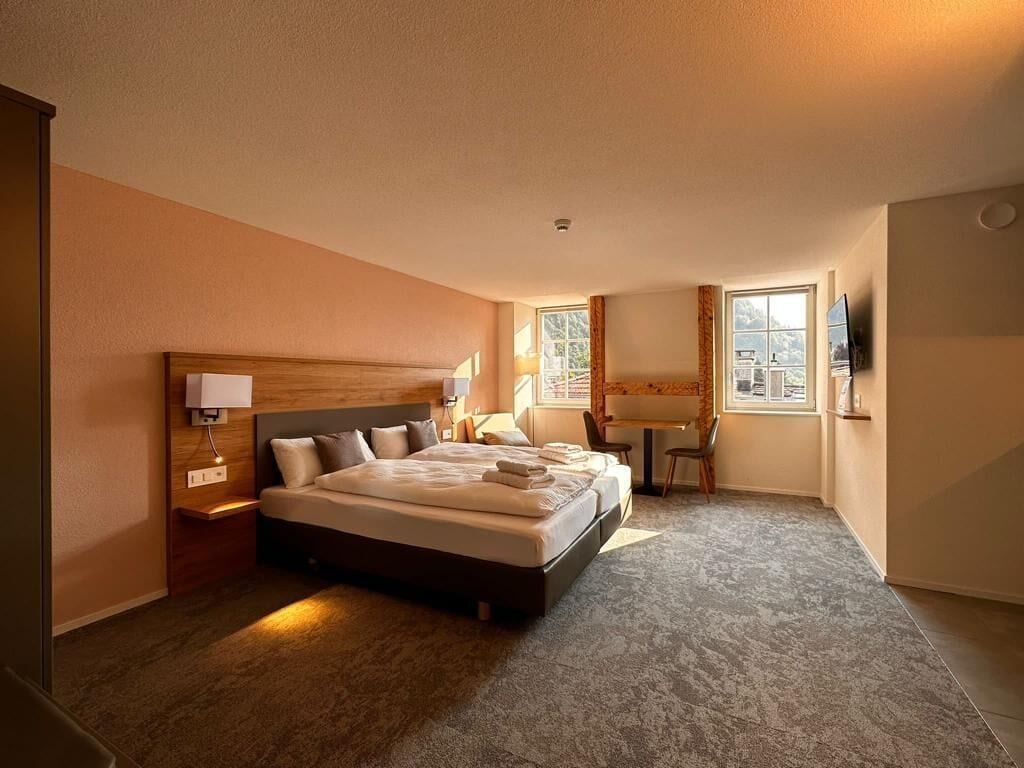 A hotel room with a large bed, a desk, and a chair. There is a window with curtains, a lamp, and a TV on the wall.