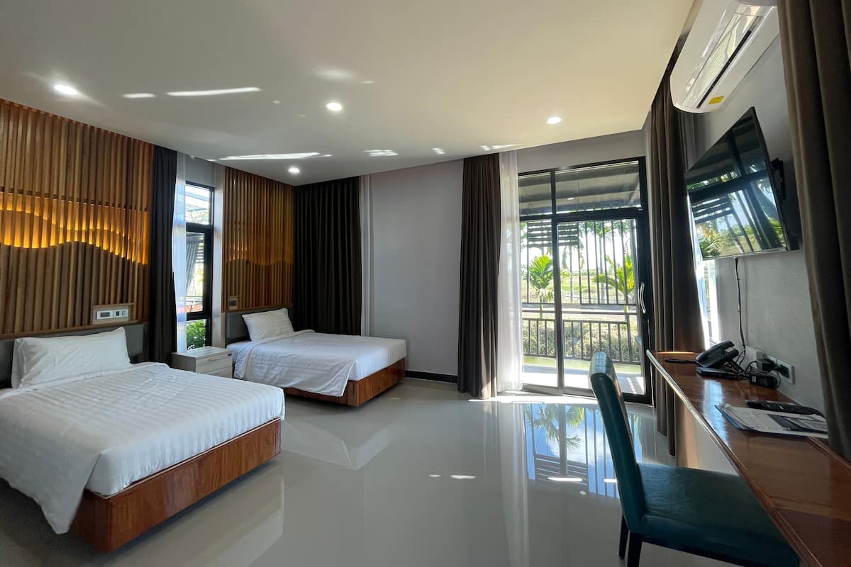 A hotel room with two beds, a desk, a chair, a TV, and a balcony with a view of greenery.