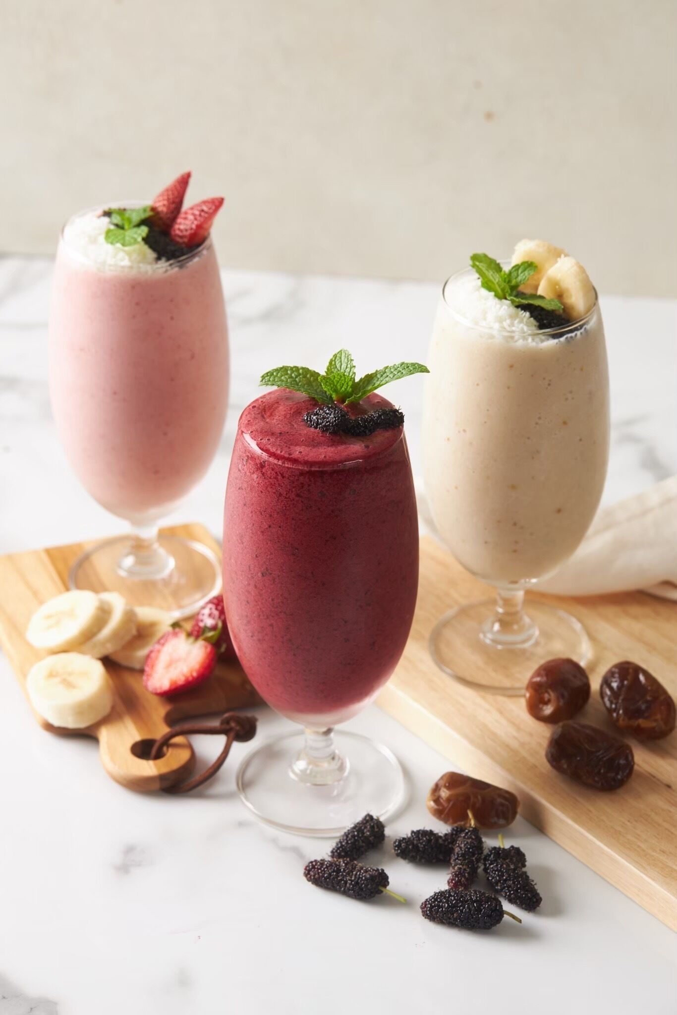 Drie smoothies in glazen, versierd met fruit en munt, op een houten plank met bananen en dadels ernaast.
