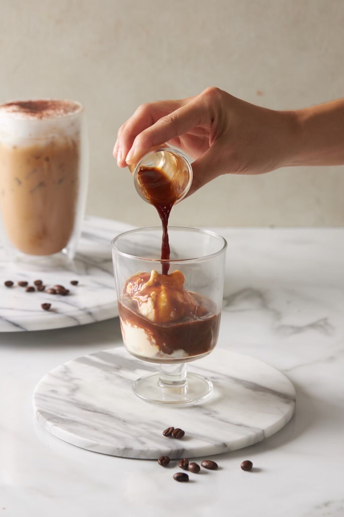 A hand pouring chocolate sauce into a glass with ice cubes and a dessert on the side.