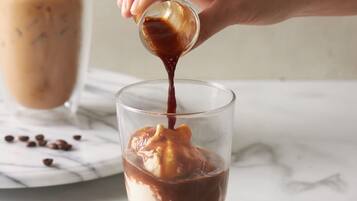 A hand pouring chocolate sauce into a glass with ice cubes and a dessert on the side.