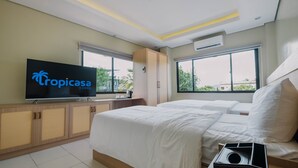 Habitación de hotel con dos camas, un televisor mostrando 'Tropicasa CORON', un armario y ventanas amplias con vistas a zonas verdes.