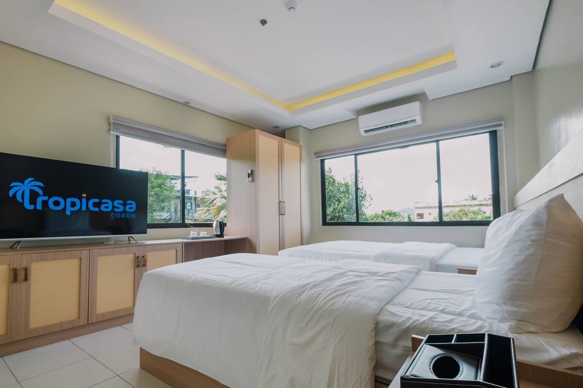 A hotel room with two beds, a TV displaying 'Tropicasa CORON', a wardrobe, and large windows with a view of greenery.