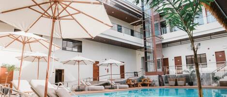 Zona de piscina con sillas de descanso y sombrillas, rodeada por un edificio de varios pisos con balcones.