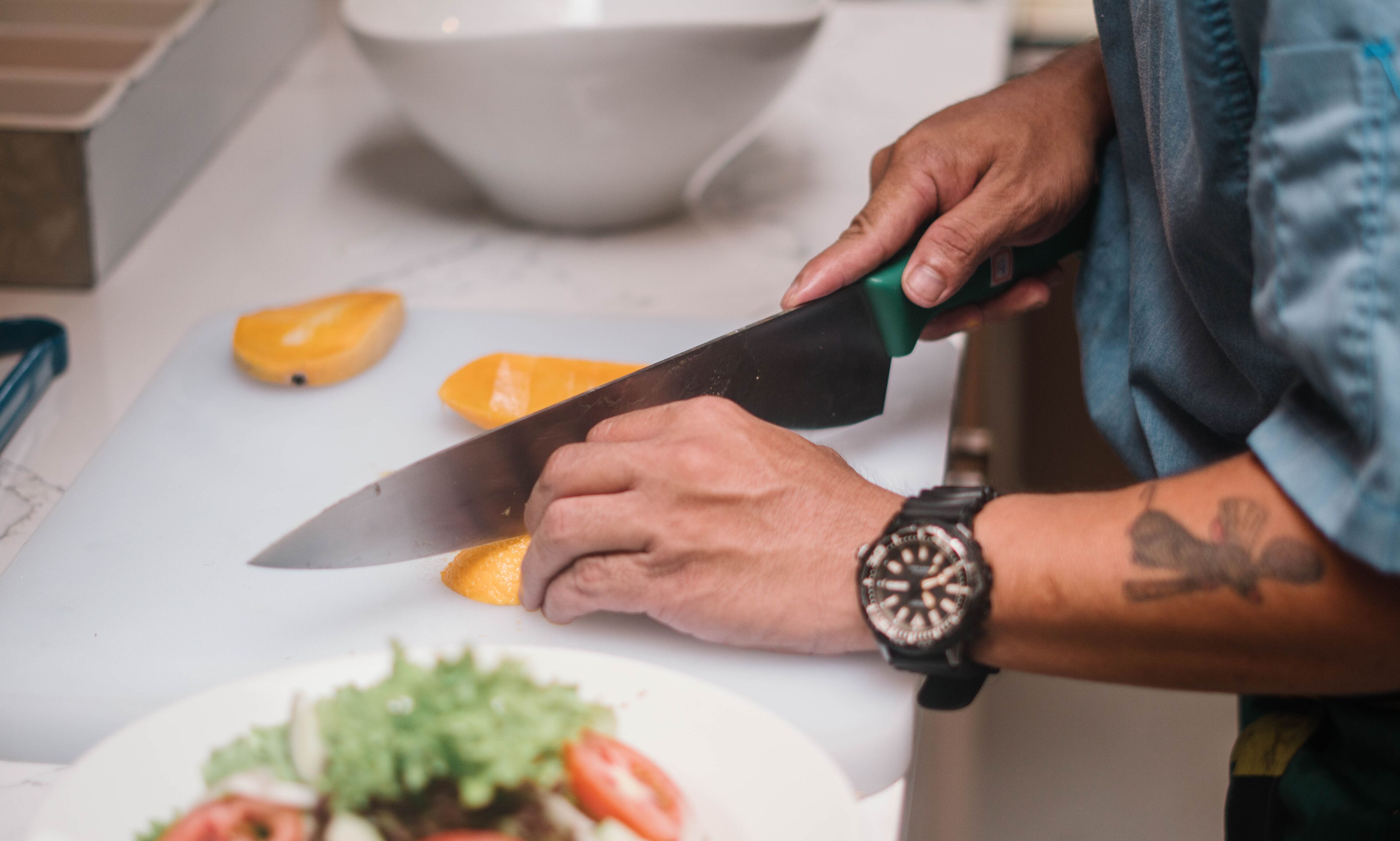 A person using a large knife to cut a fruit on a white cutting board.
