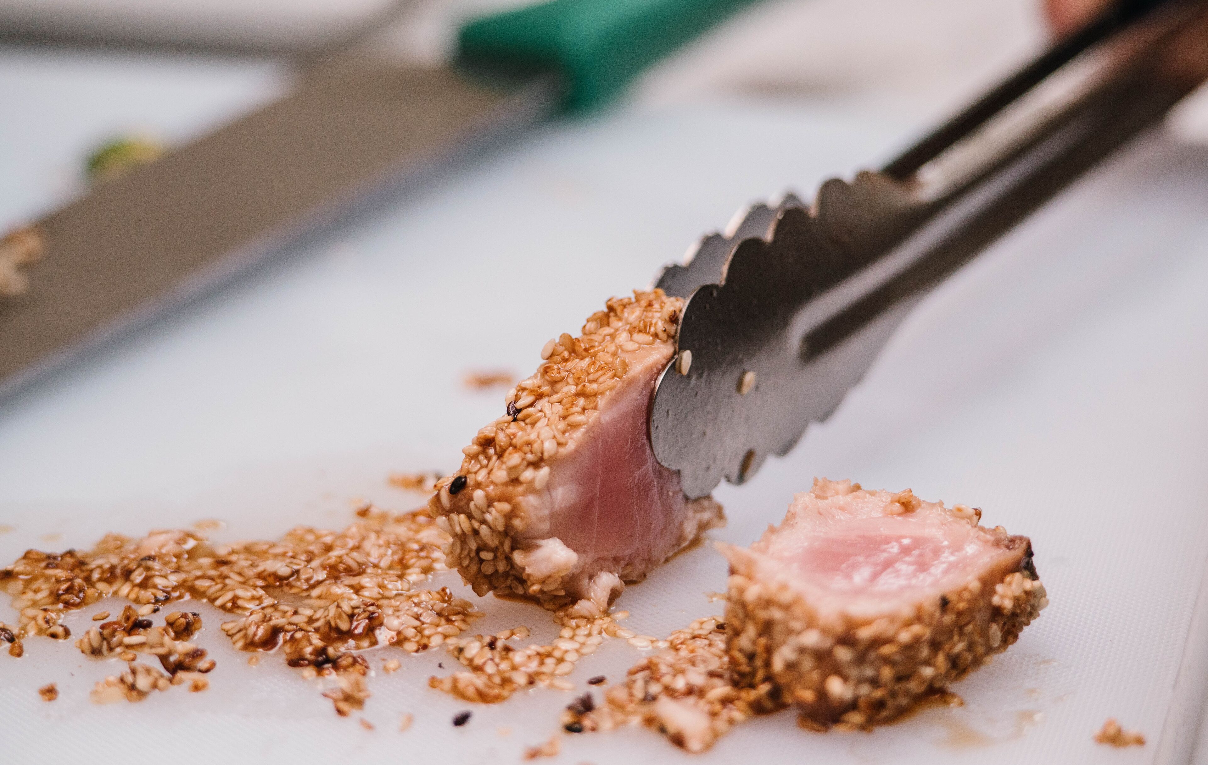 A close-up of a food preparation scene with a knife and tongs handling a piece of meat coated in sesame seeds.