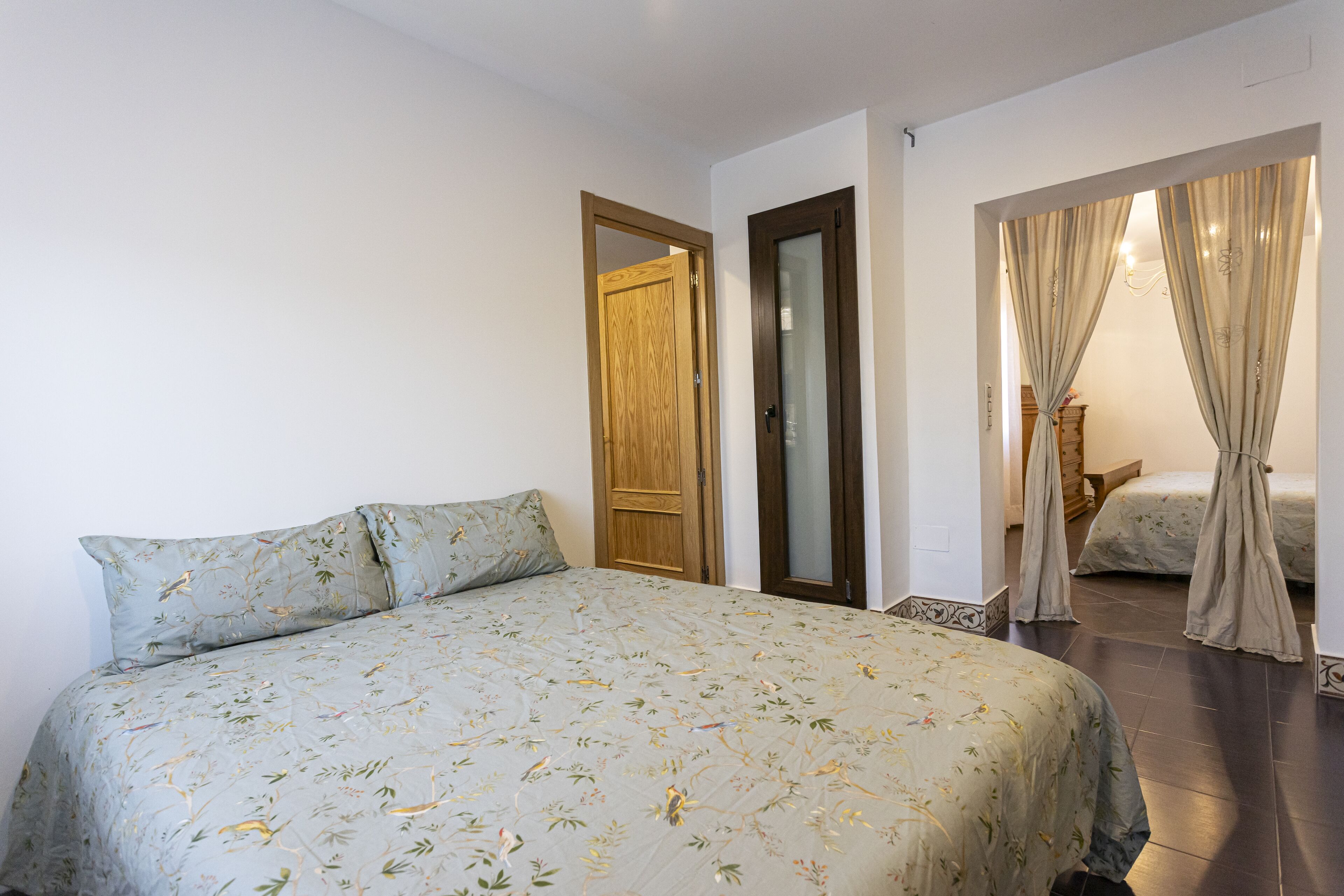 A bedroom with a bed, a wooden door, and a view through curtains to another room.