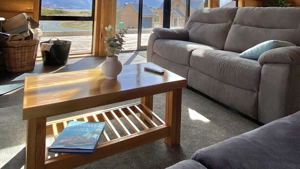 A cozy log cabin interior with a large window offering a mountain view, a comfortable sofa, and a wooden coffee table. - Log cabin - beautiful, relaxing getaway with breathtaking views! (Lake Ōhau)