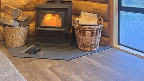A log cabin interior with a wood-burning stove, wooden walls, and a large window offering a view. - Log cabin - beautiful, relaxing getaway with breathtaking views! (Lake Ōhau)