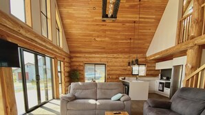 A spacious living area with a wooden ceiling, large windows, a grey sofa, a wooden coffee table, and a kitchen in the background. - Log cabin - beautiful, relaxing getaway with breathtaking views! (Lake Ōhau)