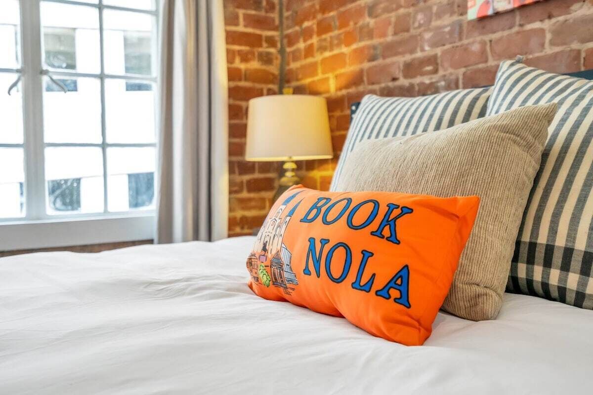 A bed with a pillow that reads "A BOOK NOLA" and a lamp on a brick wall.