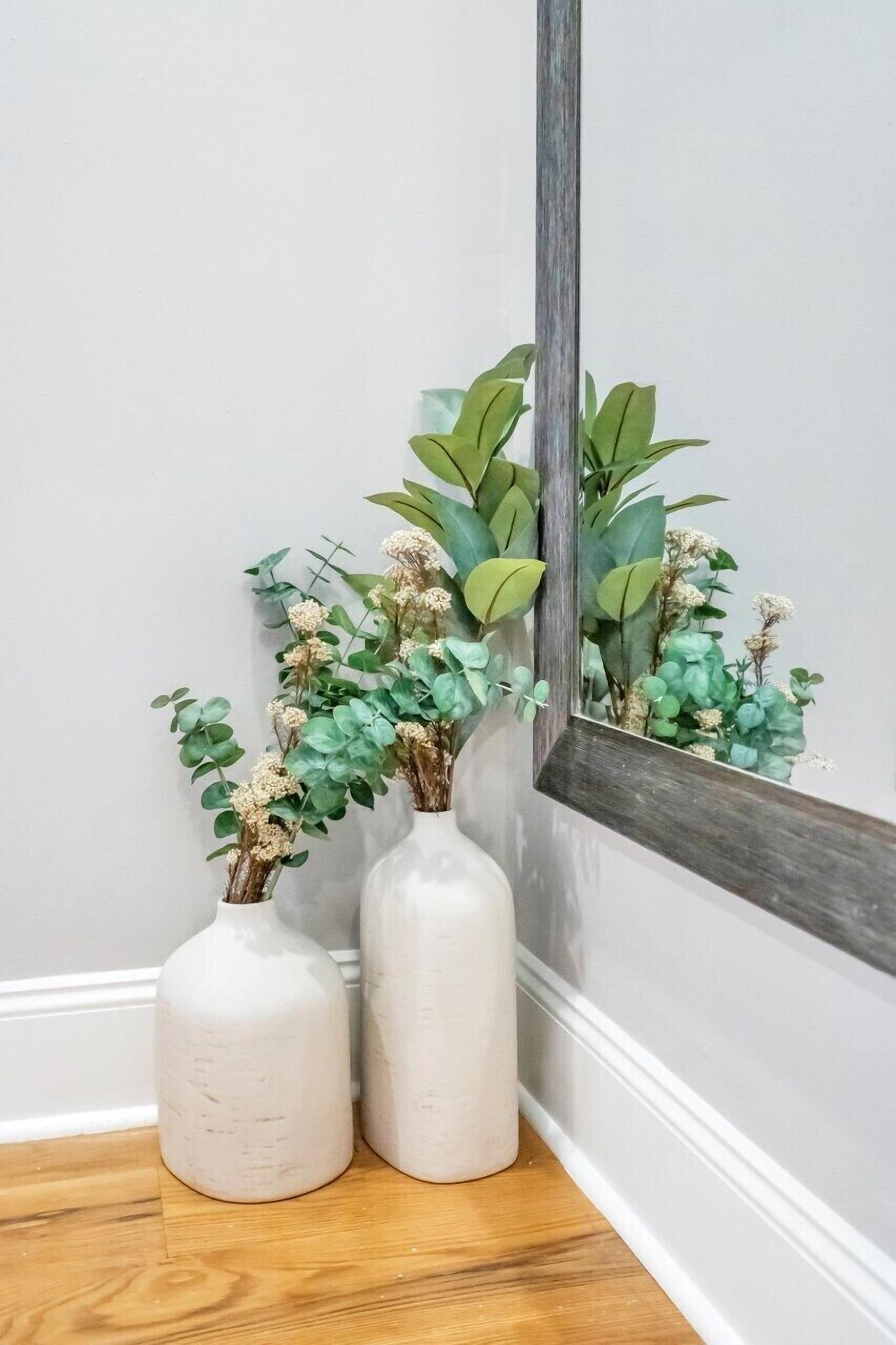 Two white ceramic vases with green foliage and dried flowers on a wooden floor against a wall with a mirror reflecting the vases.