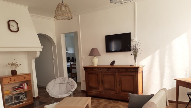 A living room with a sofa, armchair, and table, a television, and a kitchen in the background.