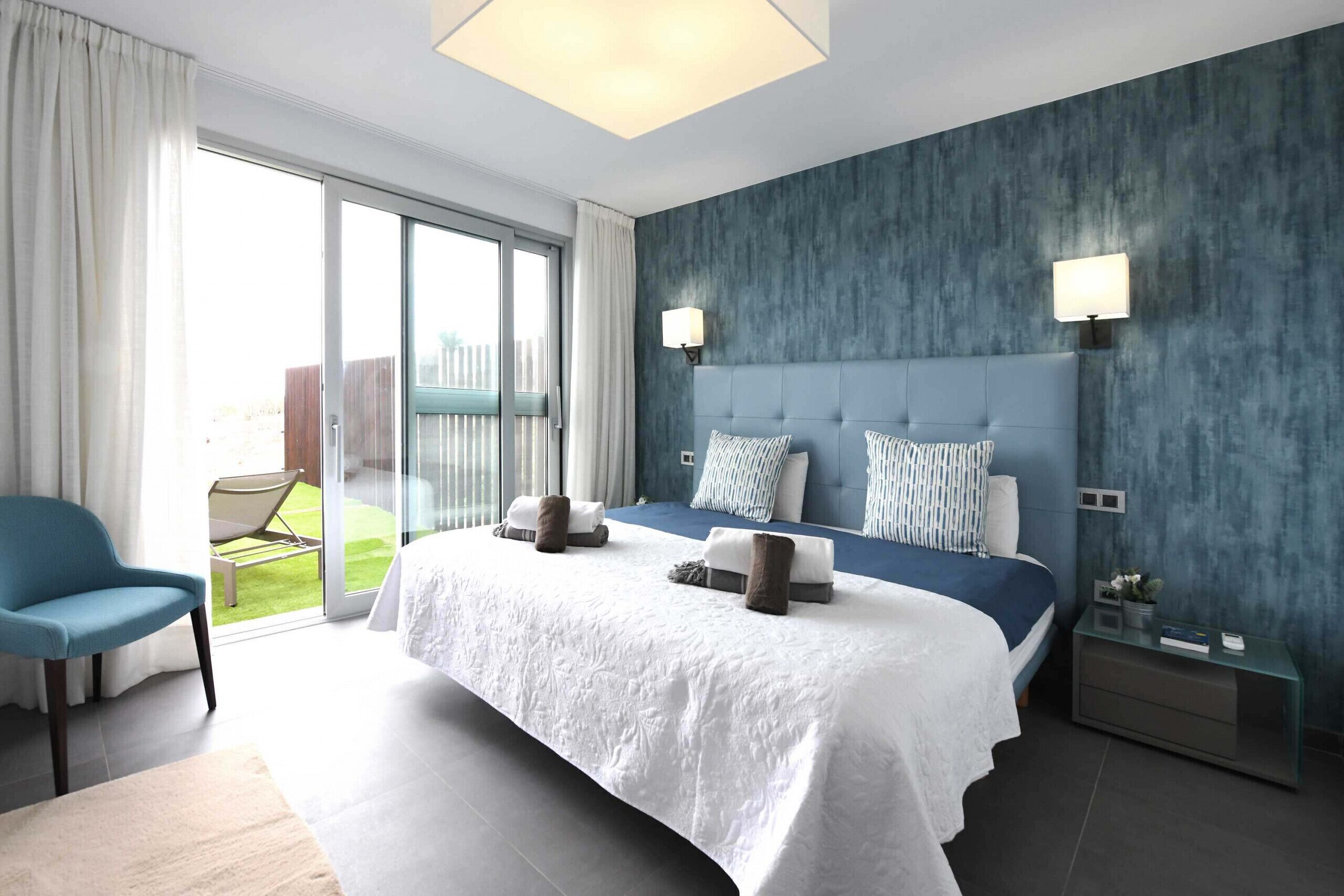Una habitación de hotel moderna con una cama grande, una silla azul, una puerta de cristal que da a un balcón y una mesita de noche con una lámpara.