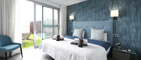 A modern hotel room with a large bed, a blue chair, a glass door leading to a balcony, and a nightstand with a lamp.