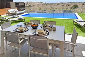A dining table set outdoors with a view of a swimming pool and a grassy area. - Salobre La Isla 7 (Maspalomas)