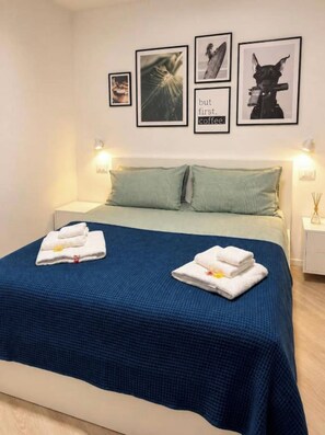 Ein Hotelzimmer mit einem Bett, zwei Kissen, einer blauen Tagesdecke, zwei gefalteten Handtüchern und einer Wand mit gerahmten Bildern.