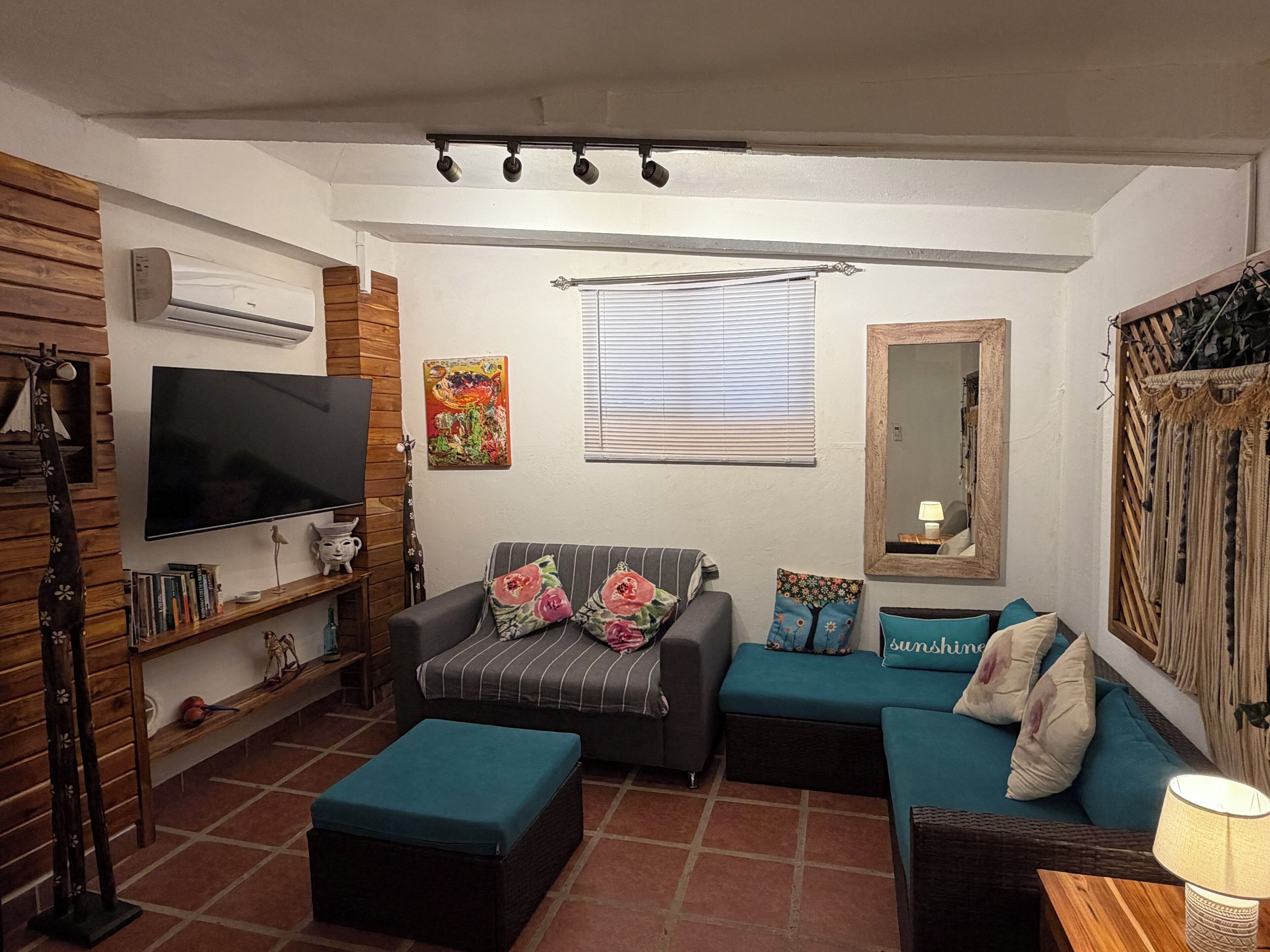 Una sala de estar con televisor de pantalla plana, un sofá, un sillón y una mesa de centro. Se observa una estantería con libros y objetos decorativos, un cuadro en la pared y una ventana con persianas.