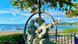 Een standbeeld van een man en een vrouw die op een bank zitten met een gitaar, omgeven door groen en met uitzicht op een strand met boten.