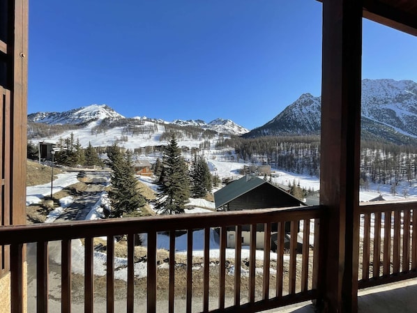 Une vue sur la montagne depuis un balcon en bois, avec des pentes enneigées et une station de ski en arrière-plan.
