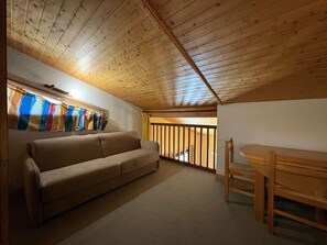 A cozy attic room with a wooden ceiling, a beige sofa, a wooden dining table, and a window with colorful curtains.