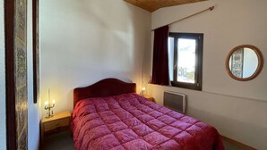 Une chambre avec un plafond en bois, un lit recouvert d’une couette rouge, une fenêtre avec des rideaux, un radiateur et un miroir fixé au mur.