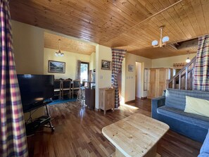 A cozy living room with wooden flooring, a television, a dining area with chairs, a sofa, and a staircase.