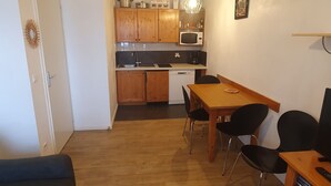 Eine kleine Küche mit Holzausstattung, einer Mikrowelle und einer Kaffeemaschine. Es gibt einen Esstisch mit Stühlen sowie einen kleinen TV-Schrank mit Fernbedienung.