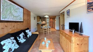 A cozy living room with a sofa, a wooden dining table with cups, and a kitchen in the background.