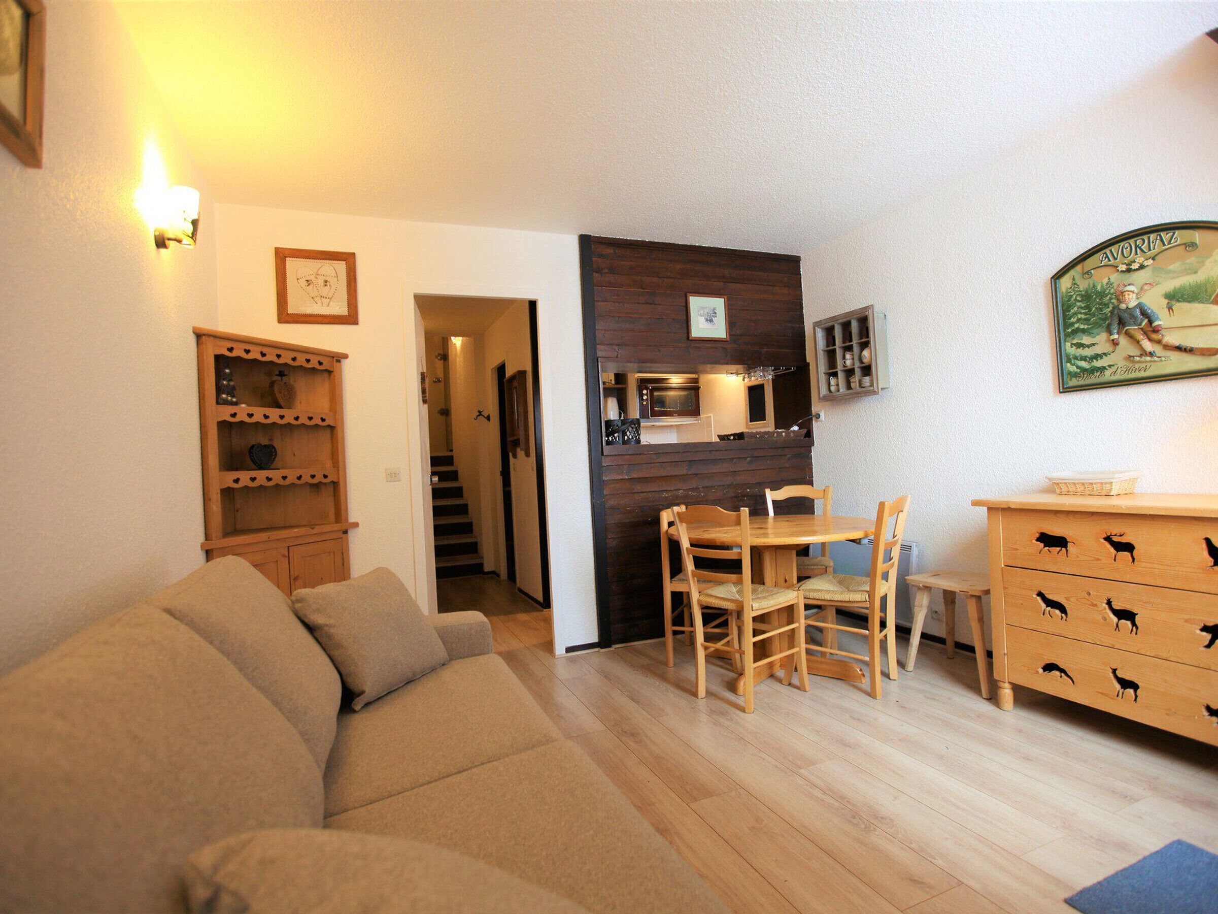 A cozy living room with a sofa, wooden dining table, chairs, a wooden cabinet with animal carvings, a kitchen area with a microwave, and a staircase leading to another room.