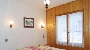 Ett sovrum med en väggpanel av trä, en säng med ett blommigt överkast, två vägglampor, en inramad tavla och ett fönster med spetsgardiner.