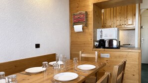 Una mesa de comedor de madera dispuesta con platos, vasos y cubiertos, con una cocina visible a través de un vano abierto.