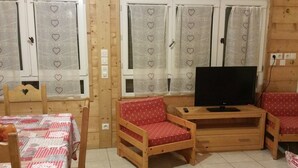 Ein gemütliches Zimmer mit Holzwänden, einem Fernseher auf einem Holzbodenständer, zwei rot gepolsterten Sesseln, einem Holztisch mit einer applizierten Tischdecke und Fenstern mit herzenmusternden Vorhängen.