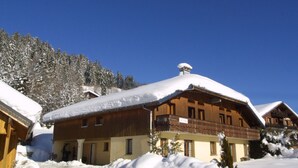 Eine Holzhütte mit schneebedecktem Dach, umgeben von Schnee, und ein bewaldeter Hügel im Hintergrund.