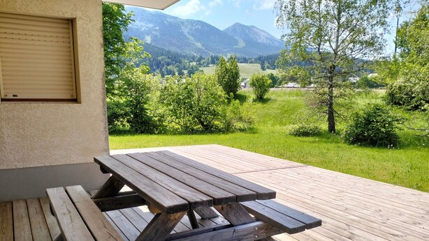Una mesa de picnic de madera con bancos en una terraza, con vistas a un paisaje montañoso cubierto de vegetación y árboles.