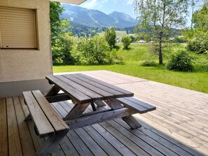 Une table de pique-nique en bois, avec des bancs, est installée sur une terrasse, d’où l’on peut admirer un paysage montagneux verdoyant, parsemé d’arbres.