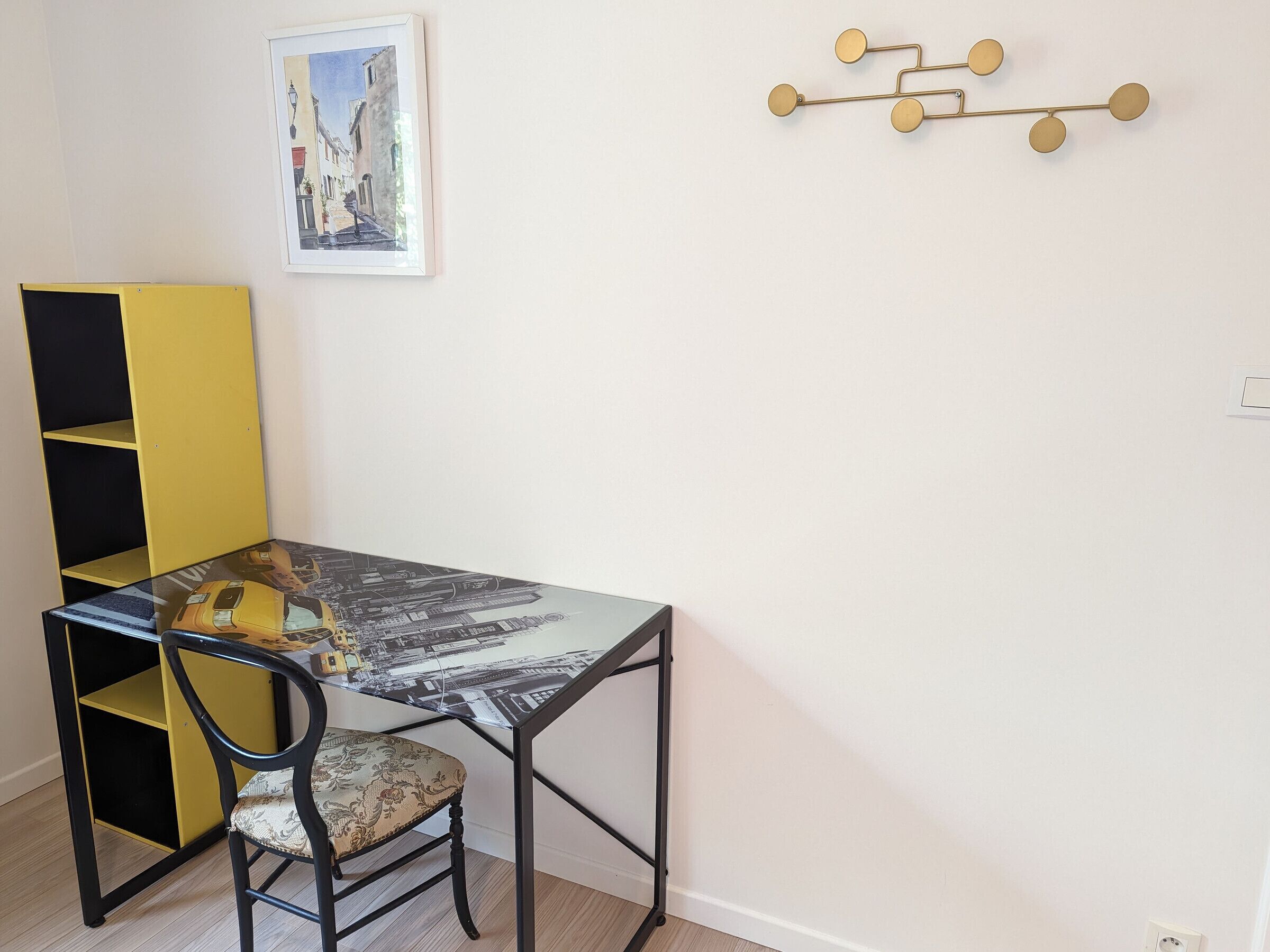 A room with a glass-top table, a yellow bookshelf, a black chair with a patterned cushion, and a wall-mounted light fixture.