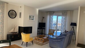 Ein Wohnzimmer mit einem Holztisch, einem gelben Sessel, einem blauen Sofa, einem Fernseher, einer Uhr und einem Fenster mit Vorhängen.
