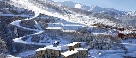 Une station de montagne enneigée, composée de plusieurs bâtiments, dont un grand chalet et de plus petites cabanes, entourée de pistes de ski et d’arbres.