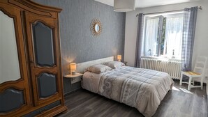 Ein Schlafzimmer mit großem Holzschrank, einem Bett mit gemusterten Betttextilien, einem Nachttisch mit Lampe, einem Fenster mit Vorhängen und einem Heizkörper.
