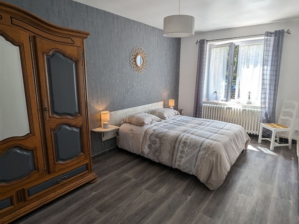 Une chambre avec une grande armoire en bois, un lit avec une literie à motifs, une table de chevet avec une lampe, une fenêtre avec des rideaux et un radiateur.