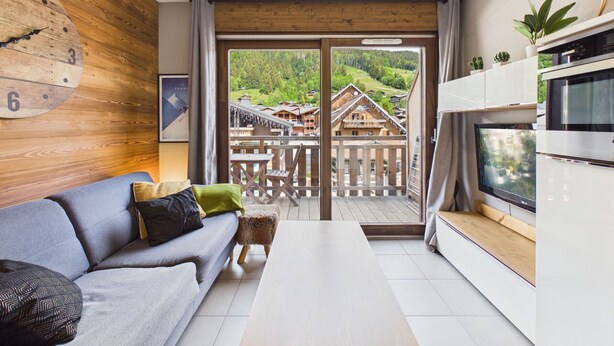 A modern living room with a sofa, a wooden wall clock, a television, and a view of a mountainous village through a glass door.