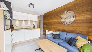 A modern kitchen with white cabinets, a wooden wall clock, a blue sofa with cushions, and a wooden coffee table.