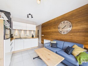 A modern kitchen with white cabinets, a wooden wall clock, a blue sofa with cushions, and a wooden coffee table.