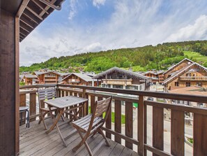 A wooden deck with a table and chairs, overlooking a village with traditional wooden buildings and a forested hill in the background.
