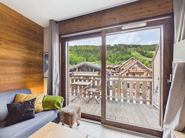 Ein Zimmer mit Holzwand, einer Couch mit Kissen, einem kleinen Hocker und einer großen Schiebeglasdoor, die auf eine Terrasse mit Holzmöbeln und Blick auf eine Berglandschaft führt.